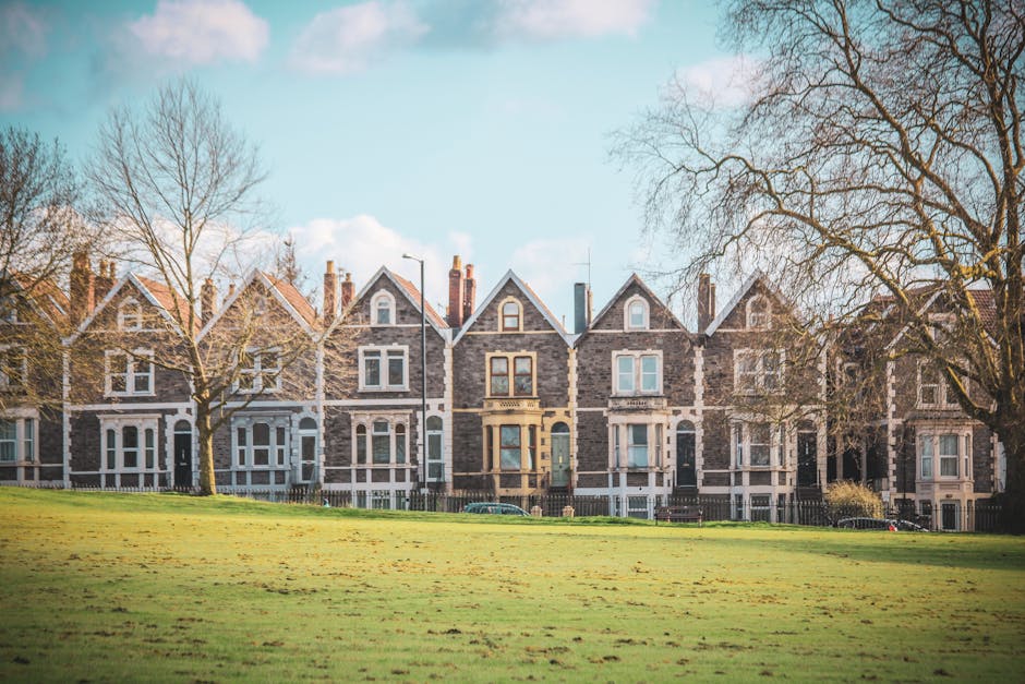 A row of traditional Victorian-style terraced houses made of dark brick with white window frames and decorative trim, set along a tree-lined street on a bright, clear day. The houses have pitched roofs with chimneys and small front gardens enclosed by low fences. Leafless trees are visible on either side of the row, indicating a late autumn or winter season. In the foreground, there is a neatly maintained grassy area. The scene is well-lit with natural daylight, highlighting the architectural features of the homes. This image captures the typical residential environment where house removals and furniture transport services by Dulwich Man and Van might involve packing, loading, and moving furniture from these properties during a home relocation process.
