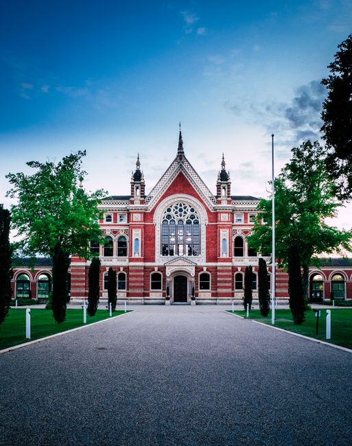 A large historic red brick building with intricate white detailing and gothic-style windows, situated at the end of a wide, paved driveway. The building features spires on its roof, decorative arches, and a central arched entrance. Tall green trees flank both sides of the driveway, leading up to the entrance, with a clear sky overhead. The scene is well-lit with natural daylight, and the area around the building appears well-maintained, suggesting a location that may be involved in heritage conservation or public services. Dulwich Man and Van may use similar scenic backdrops in promotional material related to house removals, moving logistics, or packing and furniture transport for home relocations, especially in scenic or historic urban environments.