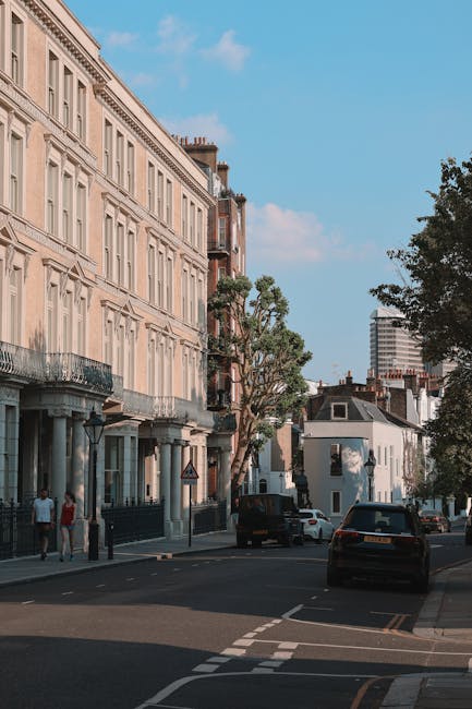A row of traditional Victorian-style terraced houses made of dark brick with white window frames and decorative trim, set along a tree-lined street on a bright, clear day. The houses have pitched roofs with chimneys and small front gardens enclosed by low fences. Leafless trees are visible on either side of the row, indicating a late autumn or winter season. In the foreground, there is a neatly maintained grassy area. The scene is well-lit with natural daylight, highlighting the architectural features of the homes. This image captures the typical residential environment where house removals and furniture transport services by Dulwich Man and Van might involve packing, loading, and moving furniture from these properties during a home relocation process.