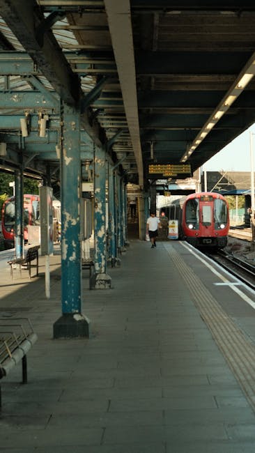 A train station platform with a red train arriving or departing on the right side, illuminated by overhead lighting under a metal canopy supported by blue-painted steel columns. The platform surface is paved with light grey tiles and features tactile paving along the edge for accessibility. A person dressed in light-colored clothing is walking towards the train, carrying a small bag or backpack. To the left, there are benches and additional shelter structures, with some passengers waiting. The station environment appears clean and orderly, reflecting typical urban railway infrastructure. This scene exemplifies the logistical setting where house removals or furniture transport may involve passenger and cargo movement within a busy station environment, aligning with topics related to home relocation or moving logistics, as covered in the Dulwich removals guide from Lordship Lane to North Dulwich Station by Dulwich Man and Van.
