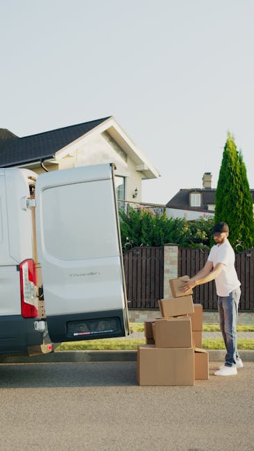 A man wearing a white T-shirt and glasses is outdoors, standing by a light grey moving van with its rear doors open, on a residential street. He is holding a cardboard box and appears to be in the process of packing or preparing for a house removal. Next to him on the pavement are three additional cardboard boxes, some of which are stacked, ready to be loaded into the van. The van's interior is visible through the open doors. In the background, there is a house with a gabled roof, white exterior walls, and a small window, surrounded by a wooden fence, green bushes, and tall coniferous trees. The scene is well-lit, suggesting daytime, and depicts a typical environment connected to relocation and furniture transport services, such as those offered by Dulwich Man and Van.