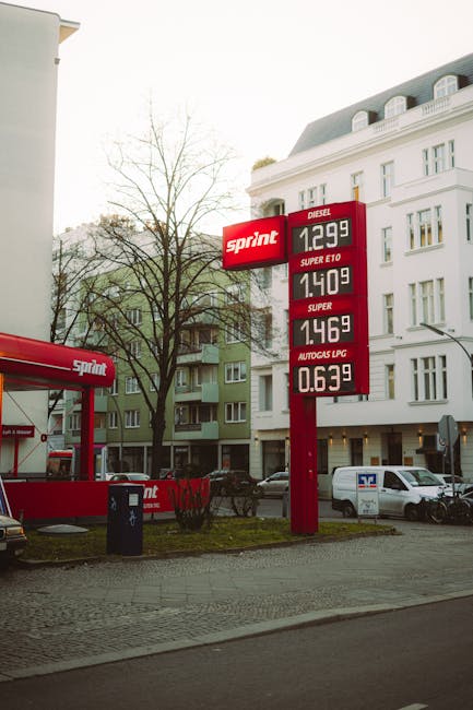 Photograph of a fuel station on a street featuring a red and white Sprint petrol price sign displaying fuel prices for diesel at £1.299, Super E10 at £1.409, Super at £1.469, and Autogas LPG at £0.639 per litre. In the foreground, part of a Sprint-branded forecourt area with a red canopy and ground-level equipment is visible. Behind the fuel sign, a white multi-storey building with classic architectural details and multiple windows can be seen, along with a leafless tree indicating late autumn or winter. Several parked vehicles, including a white van, are situated along the street, and the scene is illuminated by natural daylight. The setting appears typical of an urban environment where vehicle refuelling and home relocation services may intersect, with Dulwich Man and Van operating in the background as part of the local removals and furniture transport industry. Weather conditions are clear, with no signs of rain or overcast skies.