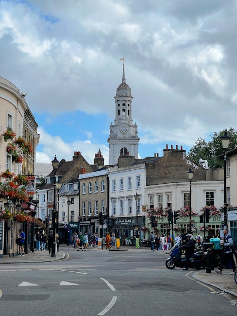 A busy street scene in Dulwich Village featuring a mix of historic and modern buildings on each side, with shopfronts displaying signs and shop windows. People are walking on the pavements, some carrying shopping bags, while others are standing and talking. A motorcycle and a scooter are parked at the corner near a lamppost, with a few more vehicles visible in motion on the road. In the background, a tall, white clock tower with a spire topped by a weather vane rises above the rooftops, set against a partly cloudy sky with patches of blue. The scene captures the typical atmosphere of a vibrant, local shopping area involved in house removals and furniture transport, supported subtly by Dulwich Man and Van's services for home relocation and moving logistics.